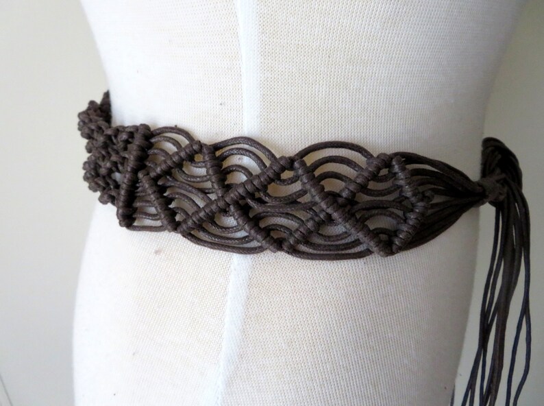 Puede incluir: Un cintur&oacute;n de macram&eacute; marr&oacute;n con un dise&ntilde;o tejido. El cintur&oacute;n se muestra en un maniqu&iacute; y presenta una serie de nudos interconectados, creando un patr&oacute;n texturizado. El cintur&oacute;n tiene un cierre de lazo con largos hilos colgantes.