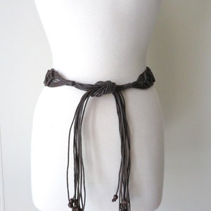 Puede incluir: Un cintur&oacute;n de macram&eacute; marr&oacute;n con un dise&ntilde;o anudado y hebras colgantes. El cintur&oacute;n presenta cuentas de madera al final de las hebras. El cintur&oacute;n se muestra en un maniqu&iacute; blanco.