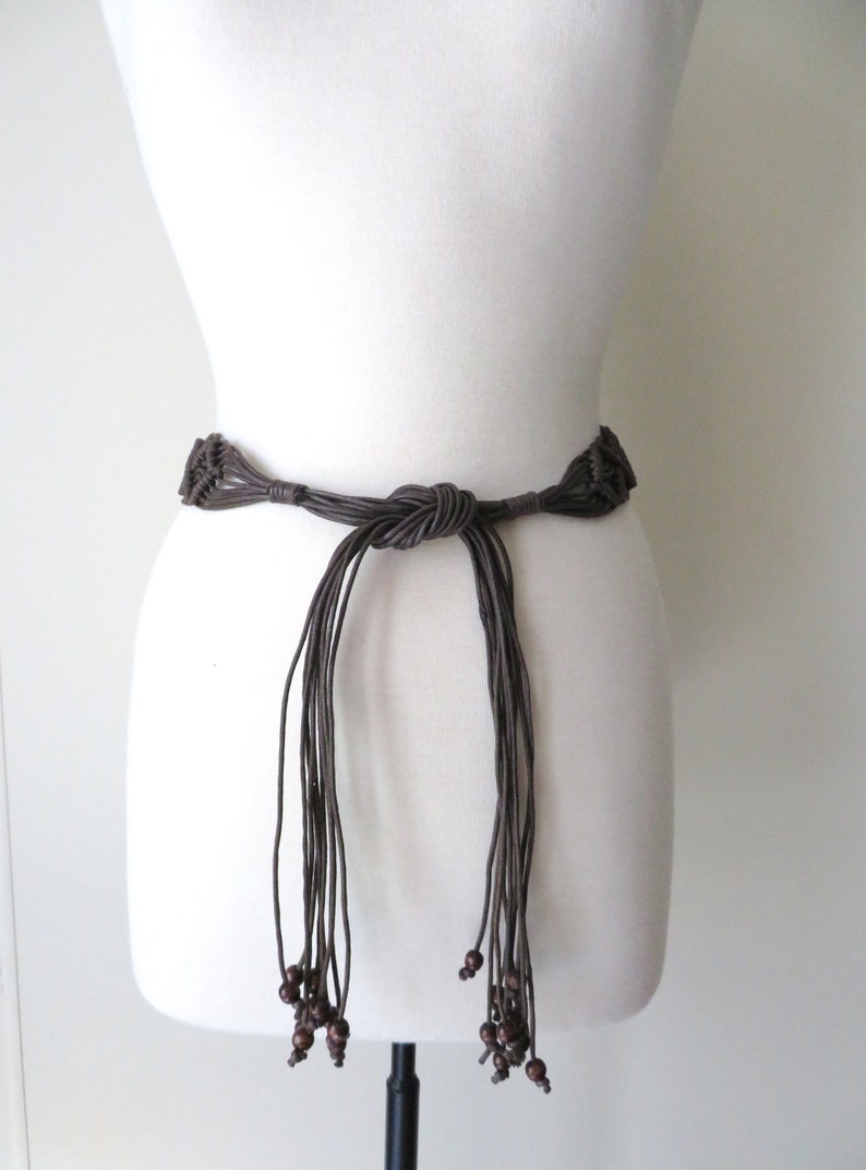 Puede incluir: Un cintur&oacute;n de macram&eacute; marr&oacute;n con un dise&ntilde;o anudado y hebras colgantes. El cintur&oacute;n presenta cuentas de madera al final de las hebras. El cintur&oacute;n se muestra en un maniqu&iacute; blanco.