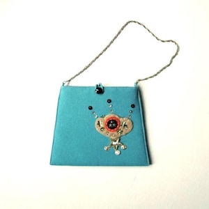 May include: A turquoise handbag with a thin silver chain strap and a black button closure. The front features a beige fabric appliqué decorated with a central orange circular element, black beads, and small silver sequins arranged in a pattern.