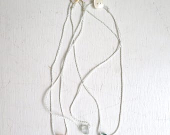 May include: Three necklaces featuring thin white braided cords, each secured with a small white button closure. Each necklace holds a single small, polished, irregular gemstone pendant in orange, clear, and teal blue colors respectively.