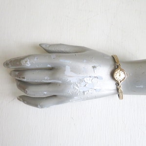 May include: A gold-tone analog wristwatch featuring a round face with a light-colored dial and Roman numeral markers. The watch has a slender, textured metal link bracelet that connects to the watch case with a decorative, open-work design.