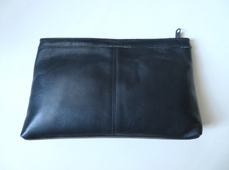 May include: A black leather clutch with a zipper closure. The clutch has a seam down the center and is made of a smooth, textured leather.