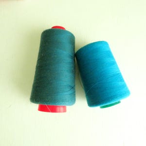 May include: Two spools of thread in shades of teal and turquoise. The larger spool has a red base, while the smaller one has a green base. The thread is tightly wound around the spools, ready for crafting projects.