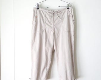May include: Beige cropped pants featuring a drawstring waist, two front pockets with button closures, and adjustable tie-string hems. The pants are displayed on a metal hanger against a plain white background, showcasing a simple design.