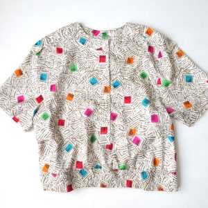 May include: A white short-sleeved button-up shirt with a colorful geometric pattern. The shirt has a round neckline and a gathered elastic waistband.