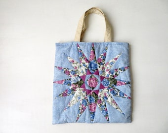May include: A blue denim tote bag featuring a floral star design. The star is made from various floral fabrics in shades of pink, blue and white.