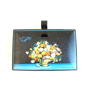 May include: Rectangular black tray with a floral arrangement in a gold vase. The tray has a black handle and a blue border. The floral arrangement includes yellow, orange, white, and blue flowers. The background has a dark swirl pattern.