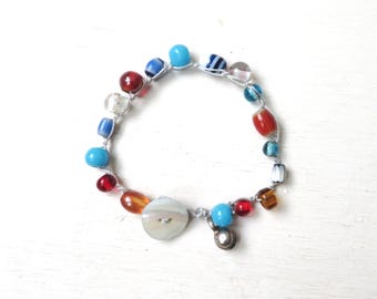 May include: A bracelet constructed from a light-colored cord featuring an assortment of multi-colored glass beads in shades of red, blue, amber, and white, along with a single round, off-white button and a small metallic charm attached.