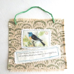 May include: A square fabric wall hanging with a beige and green scalloped pattern, featuring a green yarn hanger. A rectangular patch shows a blue and orange bird on a branch with the text "Lazuli Bunting". Below, a second patch displays the Rumi quote: "I want to sing like the birds sing, not worrying about who hears or what they think."
