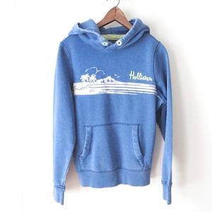May include: A blue long-sleeve hooded sweatshirt featuring a white graphic of palm trees and a mountain landscape with the text Hollister in yellow. The garment includes a front kangaroo pocket, a hood with light green eyelets, and ribbed cuffs.