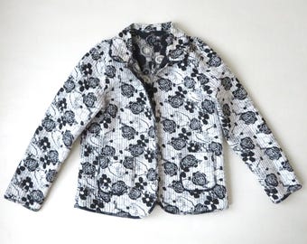 May include: A white quilted jacket with a black floral pattern. The jacket has a collar, long sleeves, and a button closure.