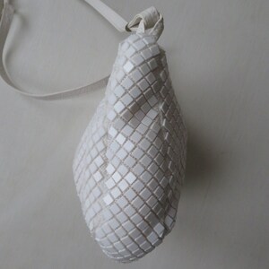 May include: A white, small, square-shaped handbag with a white strap. The bag is made of a textured material that resembles small squares.