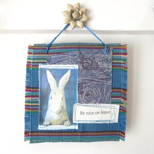 May include: A square fabric wall hanging featuring a blue striped background with frayed edges. It includes a rectangular patch with a white rabbit image and a smaller white patch with the printed text "Be nice or leave." It hangs from a blue cord.