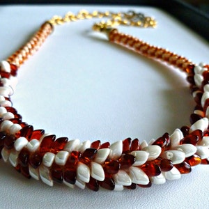 Könnte beinhalten: Eine weiße und braune Perlenkette mit geflochtenem Design. Die Kette besteht aus kleinen, runden Perlen, die in einem Spiralmuster angeordnet sind.