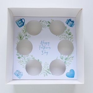 May include: White cupcake box with six circular cutouts. The interior features a design with the text "Happy Father's Day" surrounded by green leaves and blue accents, including a mug, gift, heart, and envelope.