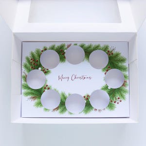 Puede incluir: Una caja de regalo blanca con una tarjeta de felicitación en su interior. La tarjeta presenta un diseño de corona navideña con agujas de pino verdes, bayas rojas y la frase "Merry Christmas". Se ven siete recortes circulares.