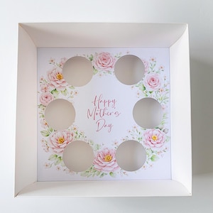 Puede incluir: Una caja blanca cuadrada con un diseño floral y el texto "Happy Mother's Day". El interior presenta una corona de rosas rosas y hojas que rodean siete recortes circulares. Ideal para regalar golosinas.