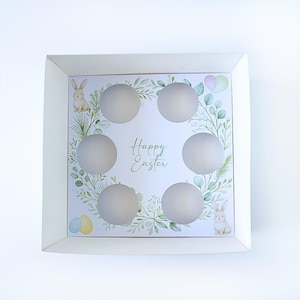 May include: White cardboard egg box with six egg-shaped recesses. The interior has a floral design with green leaves, bunnies, and the text "Happy Easter". The box is designed for Easter egg storage.