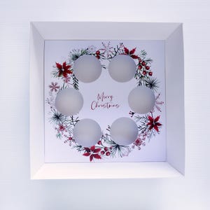 Puede incluir: Una caja blanca cuadrada con temática navideña con seis aberturas circulares. La caja presenta un diseño de corona con flores de pascua rojas, hojas verdes y copos de nieve. Las palabras "Merry Christmas" están escritas en cursiva en el centro.