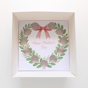 Pode incluir: Uma caixa branca com um cartão impresso no interior. O cartão apresenta um desenho de coroa de flores com folhas verdes e um laço rosa claro. O texto "Happy Mother's Day" está impresso no centro do cartão.