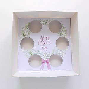 May include: White cupcake box with six circular cutouts. The interior features a floral wreath design with the text "Happy Mother's Day" in pink script, and a pink bow. The box is designed for gifting.