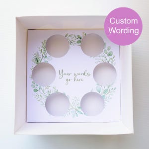May include: A white box with a custom insert featuring six circular cutouts. The insert has a wreath design with green leaves and the text "Your words go here." A pink circle with the words "Custom Wording" is in the upper right corner.