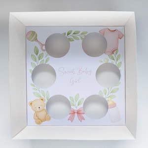 May include: White cupcake box with six circular cutouts. The interior features a pastel design with a rattle, baby clothes, a teddy bear, a bottle, and the text "Sweet Baby Girl". A pink bow is also included.