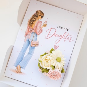 Peut inclure: Carte de vœux dans une boîte blanche avec le texte "FOR MY Daughter". La carte présente une illustration d'une femme tenant un cupcake. Un arrangement floral de fleurs en crème au beurre aux accents blancs, roses et verts orne la carte.