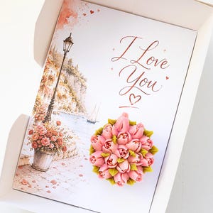 May include: A greeting card inside a white box, featuring a watercolor illustration of a coastal scene with a lamppost and flowers. The card has the words "I Love You" in elegant script, and a pink floral arrangement.