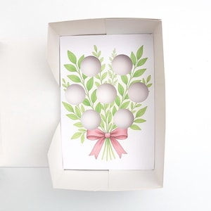 May include: A white gift box with a floral design. The card features a bouquet of green leaves and white spheres, tied with a pink bow. The box is open, revealing the card inside.