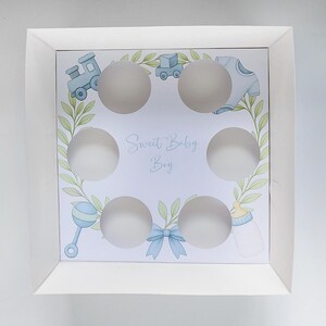 May include: White cardboard cupcake box with six circular cutouts. The interior features a light blue and green design with baby-themed illustrations, including a train, a onesie, and a baby bottle. The text "Sweet Baby Boy" is written in cursive.