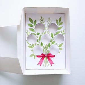 Pode incluir: Uma caixa de presente branca com um design floral. O cartão no interior apresenta um buquê em aquarela de folhas verdes e círculos brancos, amarrado com um laço rosa brilhante. A caixa está aberta, revelando o design do cartão.