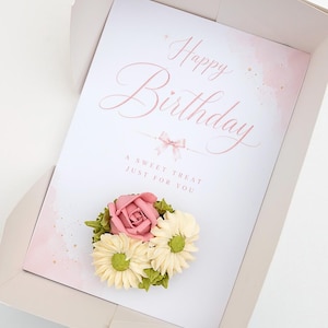 May include: A birthday card in a box with a floral arrangement. The card has the words "Happy Birthday" in pink script. The floral arrangement includes a pink rose, white daisies, and green leaves. The box is light pink.