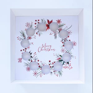 Può includere: Decorazione natalizia con cornice bianca con un design a ghirlanda con sfere di vetro smerigliato. La ghirlanda include foglie verdi, bacche rosse e fiocchi di neve. Le parole "Merry Christmas" sono scritte in corsivo.