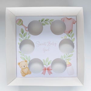 Peut inclure: Boîte à cupcakes blanche avec six découpes circulaires. L'intérieur présente un motif pastel avec un hochet, des vêtements de bébé, un ours en peluche, un biberon et le texte "Sweet Baby Girl". Un nœud rose est également inclus.
