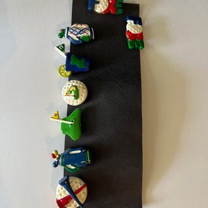 May include: A collection of golf-themed magnets on a black background. The magnets include a golf bag, golf ball, golf cart, and the word "FORE" in colorful letters. The magnets are in various colors, including green, blue, and white.