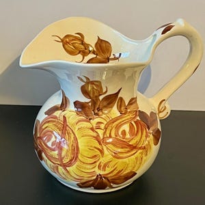 May include: Cream-colored ceramic pitcher with a wide spout and curved handle. The pitcher is decorated with yellow and brown floral designs. The handle and spout have brown accents. The pitcher is a decorative item.