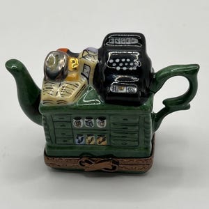 May include: A decorative teapot in the shape of a miniature office. The teapot is green with a black typewriter on top and a drawer-like base. The teapot has a brown hinged base and a green handle and spout.