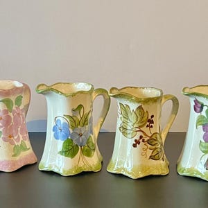 May include: Four ceramic pitchers with floral designs. Each pitcher has a unique hand-painted design, including pink flowers, blue flowers, green leaves, and purple grapes. The pitchers have a cream-colored base and green and pink accents.