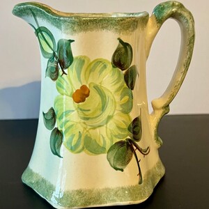 May include: Cream-colored ceramic pitcher with a large yellow and green floral design. The pitcher has a green rim and handle, with additional green floral accents. The handle is curved and the pitcher has a flared base.