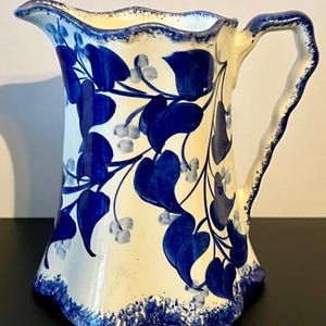 May include: A white ceramic pitcher with a blue floral design. The pitcher features a curved handle and a scalloped rim. The blue design includes leaves and berries. The pitcher is likely used for serving beverages.