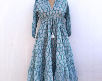 May include: A teal and white patterned dress with long sleeves and a tiered skirt. The dress features a V-neckline, a drawstring waist, and a white tassel. The dress is displayed on a mannequin.