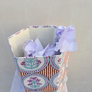 Puede incluir: Una papelera hexagonal con un patrón repetitivo de motivos florales morados y azul turquesa dentro de marcos circulares, sobre un fondo de rayas verticales naranjas y blancas. El interior contiene hojas de papel blanco arrugadas.