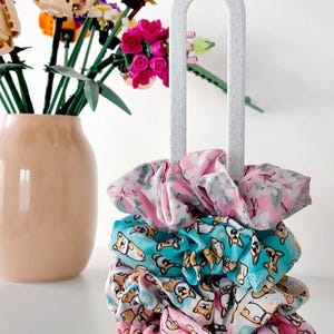 Könnte beinhalten: Ein Stapel bunter Haargummis mit Blumen- und Corgi-Mustern hängt an einem hellgrauen Halter. Im Hintergrund befindet sich eine pfirsichfarbene Vase mit einem Strauß bunter Blumen. Die Haargummis sind rosa, blau und weiß.