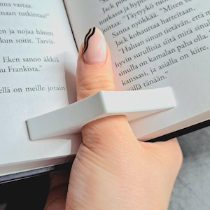 May include: A white rectangular book page holder is shown, inserted between the pages of a book. The holder is designed to keep the book open. The image is a close-up, showing the holder in use.