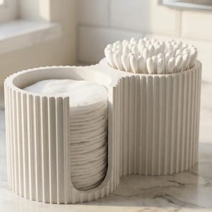Può includere: Un organizer da bagno bianco e scanalato con due scomparti. Un lato contiene una pila di dischetti di cotone rotondi, l'altro bastoncini di cotone. L'organizer è appoggiato su un piano di lavoro in marmo, con una finestra e un muro piastrellato sullo sfondo.