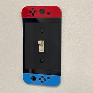 May include: A decorative light switch cover designed to resemble a video game controller. The cover features a red top and blue bottom, with black center panel. The design includes button details and a toggle switch.