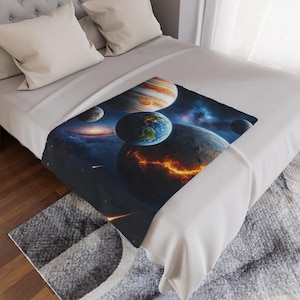 Peut inclure: Une couverture ornementale représentant des planètes et des galaxies dans l'espace. Le motif comprend la Terre, Jupiter et d'autres corps célestes sur un fond cosmique bleu foncé et violet. La couverture est présentée sur un lit avec une literie de couleur neutre.