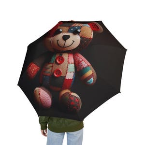 May include: Black umbrella featuring a patchwork teddy bear design. The bear has a smiling face, button eyes, and red button accents. The bear's body is made of various fabric patches in red, blue, and brown.